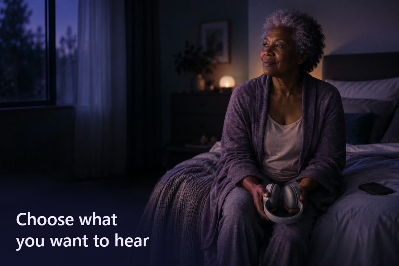 An older woman sits on the edge of a bed holding headphones in her lap, with a dark phone beside her.
