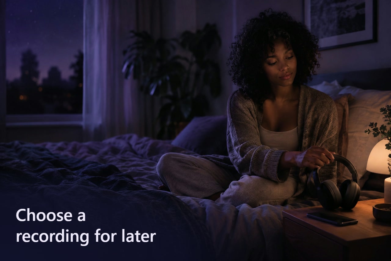A woman seated on a bed lifts over-ear headphones beside a phone on the bedside table.