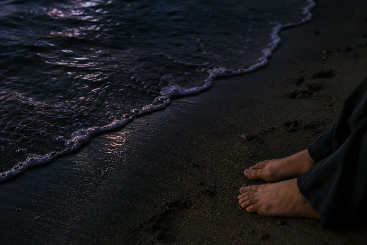 A small wave thins across wet sand near an adult's bare feet at twilight.