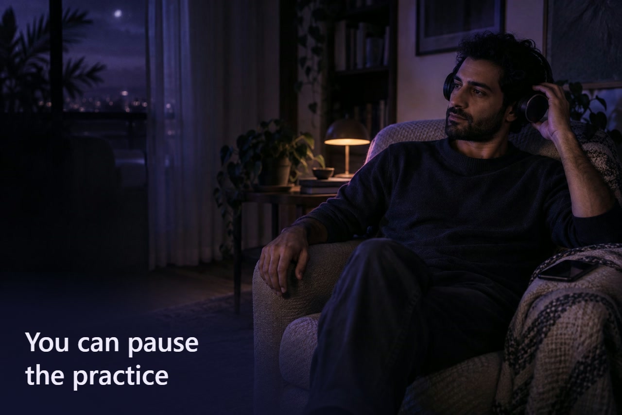 A man lifts one headphone earcup while seated in an armchair, with a dark phone beside him.